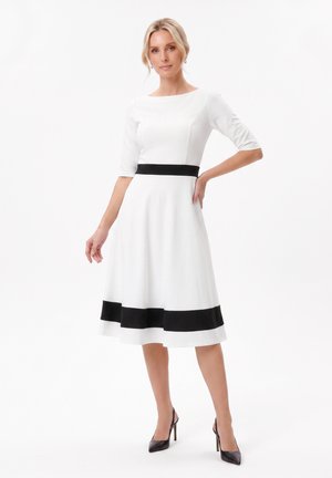 Femme debout en robe blanche longueur genou avec ceinture noire et détails sur l'ourlet, portant des talons hauts noirs et des boucles d'oreilles argentées sur fond blanc.