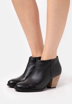 Botas de tobillo de cuero negro con un acabado texturizado, puntera puntiaguda y un tacón de madera apilada. El diseño es elegante, sin herrajes visibles.