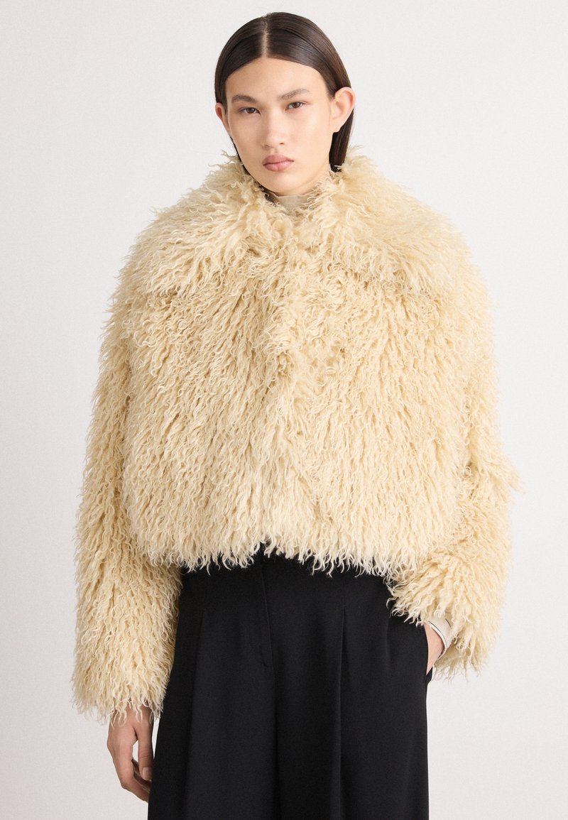 Crème-kleurige nepbont cropped jas met lange mouwen, harige structuur, ronde kraag en rechte zoom, gestyled over zwarte broeken.