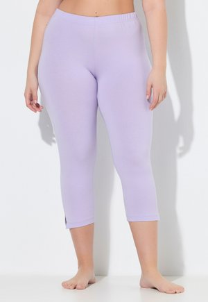 Frau trägt hellviolette kurze Leggings mit elastischem Bund, steht barfuß vor weißem Hintergrund.