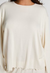 Losse crème pullover top met een geribbelde textuur en subtiele verticale lijnen, een ronde halslijn en golvende zoomranden.
