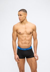 Μυώδης άντρας που φοράει μαύρα εσώρουχα Calvin Klein με μπλε μέση, κοιτάζοντας αριστερά του μπροστά σε απλό λευκό φόντο.