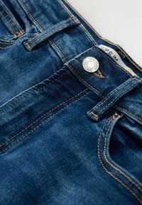 Modré džínsy z denimu s medenými nitmi, kontrastným prešívaním, kovovým gombíkom s nápisom "NEXT DENIM SINCE 1982" a viditeľnou vnútornou etiketu s nápisom "COOP SLIM".