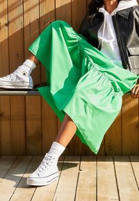 Zielona marszczona spódnica z lekkiego materiału, zestawiona z białymi sneakersami typu high-top i białymi skarpetkami, na tle drewnianej ściany.