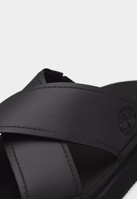 Μαύρο δερμάτινο σανδάλι με φαρδιές διασταυρούμενες λουράκια και ανάγλυφο λογότυπο δέντρου Timberland στο πλάι του λουριού, σε λευκό φόντο.