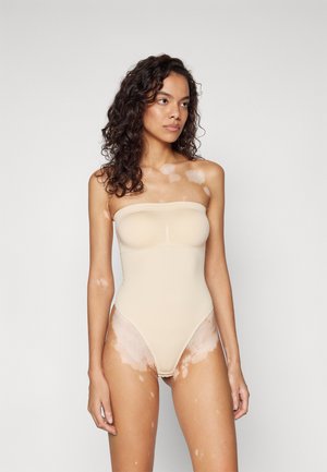 Beige stroppeløs body med en glatt tekstur, høyt kuttede ben og sømdetaljer langs brystet og hoftene.