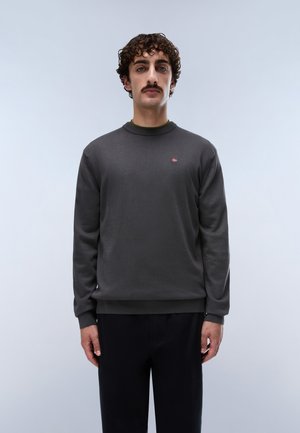 Mann mit lockigem Haar und Schnurrbart trägt dunkelgrauen Pullover mit kleinem rot-weißem Logo auf der Brust, steht vor hellem einfarbigem Hintergrund.