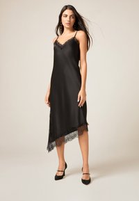 Svart satinklänning med spetskant längs den asymmetriska fållen. Har tunna band och en v-ringning för en elegant och flödande silhuett.