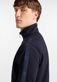 Mornarica zip-up dukserica s rebrastom teksturom, visokim ovratnikom i izvezenim logotipom na ramenu. Dizajn sadrži elegantnu, prilagođenu siluetu.