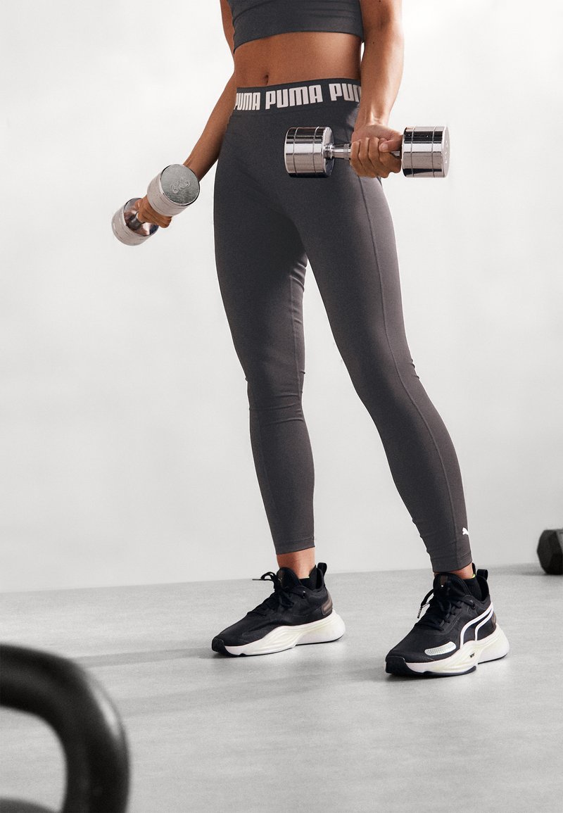 Black and gray workout outfit featuring a cropped top and high-waisted leggings with a logo band, holding chrome dumbbells, and wearing black sneakers.