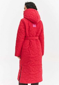 Czerwony pikowany długi płaszcz z kapturem, z fakturą w liście i paskiem w talii, prezentowany od tyłu.