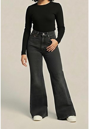 Persoon die een zwarte top met lange mouwen draagt, hooggesneden donkere wijde jeans en witte schoenen, staand tegen een effen achtergrond.