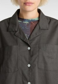 Chemise noire et grise en tissu lisse avec un col pointu, deux poches poitrine et un petit logo sur la poche droite.