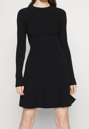 Femme portant une robe en maille noire cintrée à manches longues, avec un col rond, une jupe évasée et des manches côtelées sur fond uni.