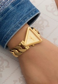Gold triangular watch with a Guess logo, featuring a linked bracelet design. Blue denim cuff partially visible, neutral skin tone beneath.