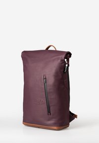 Zaino a rotolo in pelle bordeaux scuro con texture liscia; presenta una tasca laterale con zip e una base in pelle marrone.