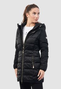 Giacca piumino nera con cappuccio, texture trapuntata, zip frontale, due tasche laterali e dettagli dorati. Indossata sopra una camicia bianca con un logo.