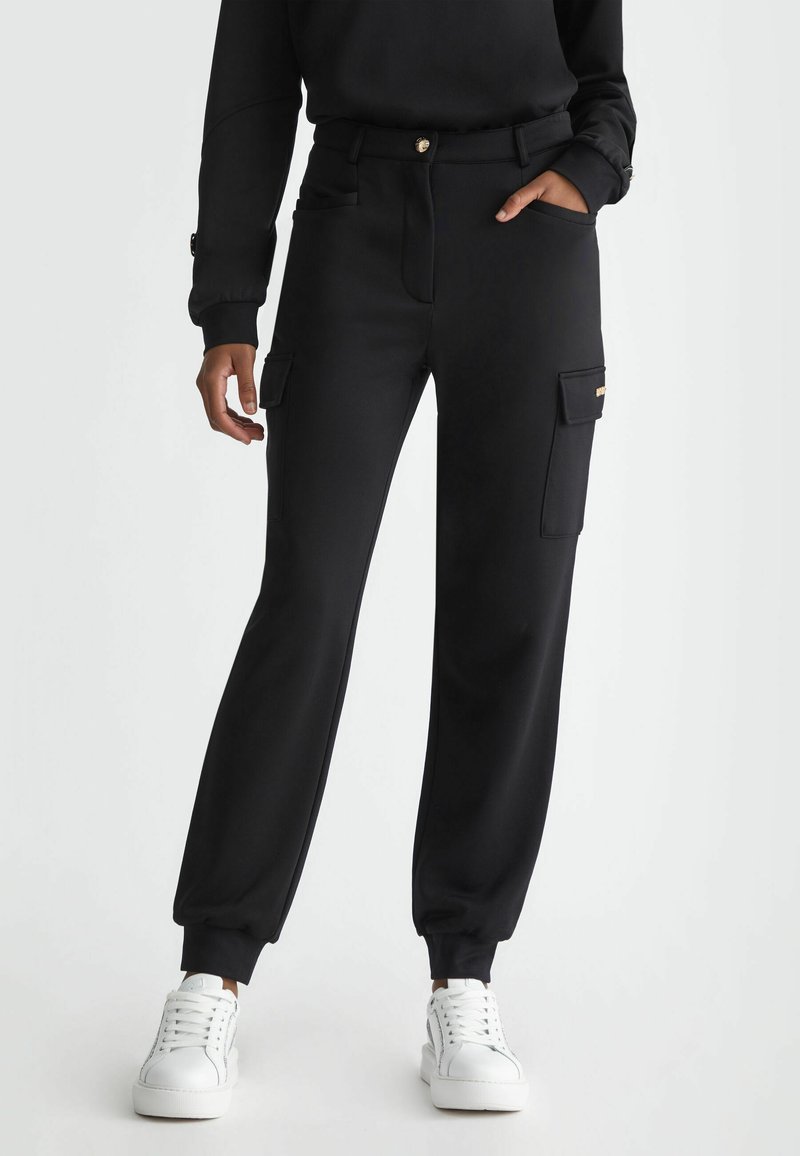 Personne portant un pantalon jogger cargo noir avec des poches latérales et des baskets blanches, debout avec une main dans la poche sur un fond uni.