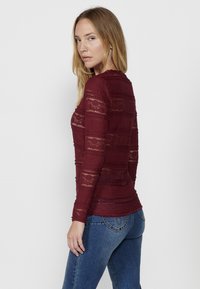 Maglietta a maniche lunghe bordeaux con dettagli in pizzo orizzontali, tessuto testurizzato, scollo rotondo e design aderente, abbinata a jeans blu.
