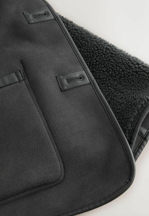 Czarny rzut na faux skórze z teksturowaną powierzchnią, z miękkim, ciemnym wnętrzem z polaru i dwoma pętlami do zapięcia. Krawędzie wykończone skórą.