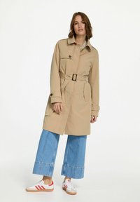 Beige trenchcoat met een ceinture taille, knoopsluiting aan de voorkant en zijzakken, gecombineerd met wijdnekkige blauwe denim jeans en witte sneakers met rode accenten.