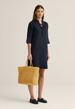 Kvinde iført en navy knælængde kjole, der holder en stor gul flettet taske, stående mod en ensfarvet beige baggrund.