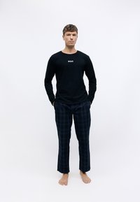 Melna garās piedurknes krekls ar "BOSS" logo; tumši zili rūtaini pidžamas bikses. Audums izskatās mīksts, un piegriezums ir ikdienišķs. Kails modelis stāv centrā.