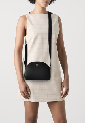 Borsa a spalla nera in pelle con forma curvata, hardware dorato e tracolla nera regolabile. Indossata sopra un vestito a quadri chiari.
