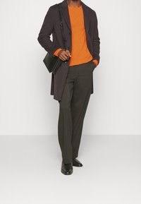 Tummanvioletti takki oranssin villapaidan päällä, yhdistetty tummanharmaisiin housuihin ja mustiin kenkiin. Musta laukku roikkuu olkapään yli.