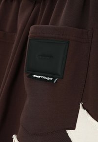 Tejido marrón con un bolsillo que presenta una placa rectangular negra. Incluye una etiqueta blanca con "MHM Design". Acento texturizado en beige contrastante.