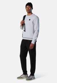 Sudadera gris claro con un logo cuadrado, combinada con pantalones jogger negros que tienen bolsillos. Usada con un gorro negro y zapatillas gris/verde.