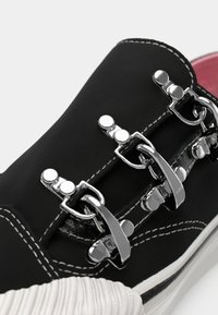 Zapato negro con parte superior texturizada, herrajes de cierre plateados, costuras en blanco en contraste y suela blanca estriada. Forrado en rosa.
