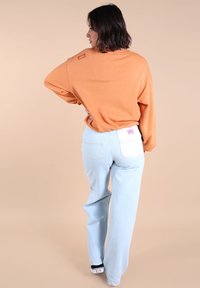 Sweatshirt orange à coupe décontractée, associé à un jean en denim clair à jambes larges avec une poche plaquée blanche et une étiquette violette. Chaussures noires.