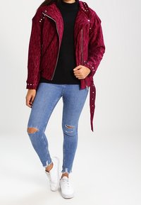 Vrouw draagt een gestructureerd bordeauxrood jasje met studs, gescheurde lichtblauwe skinny jeans, een zwarte trui en witte sneakers, staand op een witte achtergrond.