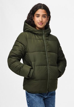 Mujer con cabello rizado oscuro que lleva una chaqueta acolchada verde oliva con capucha y vaqueros azules, de pie con las manos en los bolsillos sobre un fondo blanco.