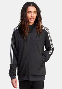 Chaqueta negra con cremallera hecha de tela suave, con acentos en blanco de tres rayas en las mangas y un logo bordado en el pecho.