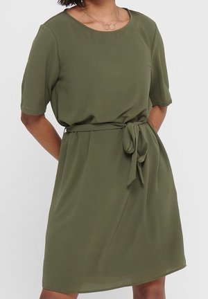 Mujer con un vestido verde oliva hasta la rodilla, de mangas cortas y cinturón anudado en la cintura, además de un delicado collar con tres anillos entrelazados.