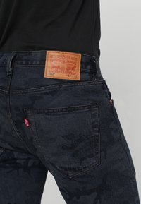 Mörkblå denimjeans med ett kamouflage-mönster, läderlogotyp vid midjan och bakfickor med röd tab-accent.
