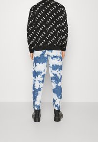 Sudadera negra con patrón de logotipo blanco, combinada con pantalones azul tie-dye. Las botas negras completan el conjunto.