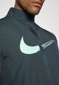 Nike-takki tummanharmaana, jossa on korkea kaulus, etuvetoketju ja suuri mintunvihreä swoosh-logo. Sujuva, venyvä kangas.
