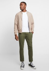 Beige bomberjakke over en hvid t-shirt, parret med olivengrønne slim-fit bukser og sorte sneakers med hvide såler.