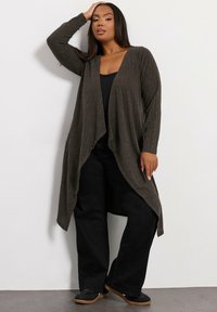 Lång öppen cardigan i strukturerat mörkt olivgrönt tyg, kombinerad med en svart topp och vida svarta byxor. Har en flödande design.