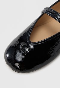 Fekete lakkbőr balettlapos, lekerekített orral, V alakú kivágással és apró díszítőelemmel az orrán.