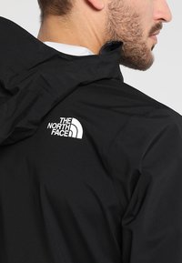 Homme portant une veste noire North Face avec capuche, cadré pour montrer la barbe, l'oreille et le logo sur le haut du dos sur un fond uni.