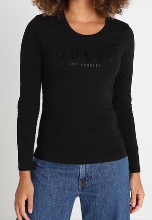 Woman wearing a black long-sleeve shirt with "GUESS Los Angeles" text and blue jeans, standing against a plain light background.