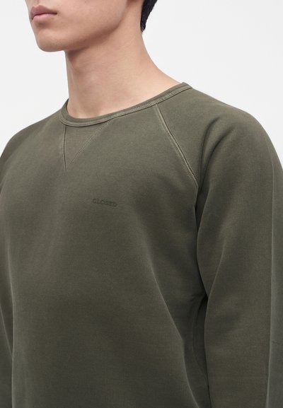 Gros plan d’une personne portant un sweat-shirt vert olive à col rond avec des coutures visibles et un logo brodé discret sur la poitrine.