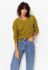 Pull en maille côtelée vert olive avec un col en V, associe à un jean en denim bleu taille haute, présentant une coupe décontractée et des manches retroussées.