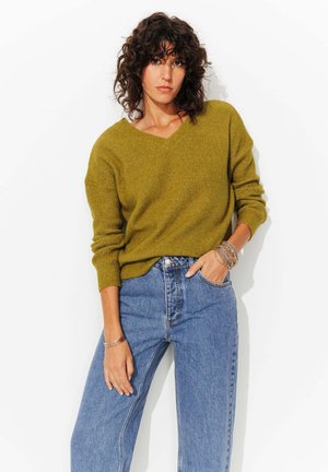 Pull en maille côtelée vert olive avec un col en V, associe à un jean en denim bleu taille haute, présentant une coupe décontractée et des manches retroussées.