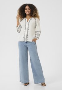 Blusa bianca con ricami neri lungo le cuciture e le maniche, dotata di scollatura a V e maniche tre quarti, abbinata a jeans a gamba larga blu chiaro.