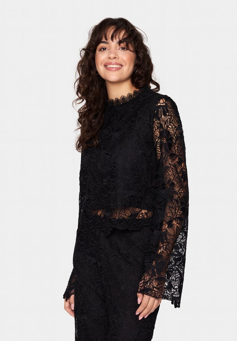 Top in pizzo nero con maniche lunghe a fantasia e scollatura alta. Presenta motivi floreali e un taglio corto. Tessuto trasparente e strutturato.